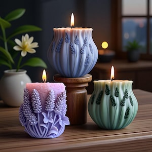 May include: Three decorative candles with lit flames, in blue, pink, and green, each with a floral design. The blue candle sits on a wooden stand. The candles have a wavy top edge. The candles are on a wooden surface.