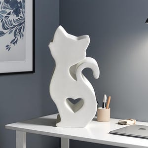 Puede incluir: Una escultura decorativa blanca con forma de gato y un recorte en forma de corazón. La escultura tiene una cola curvada y está colocada sobre una superficie blanca. El fondo incluye una impresión floral enmarcada y un portalápices.