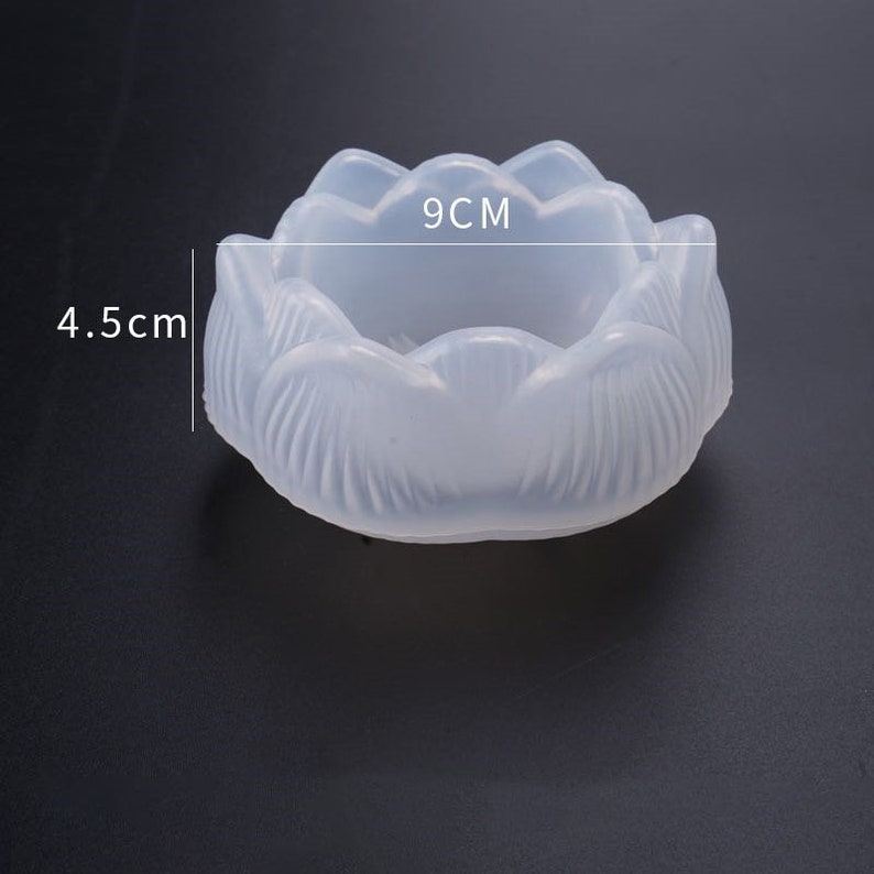 Puede incluir: Un molde de silicona transl&uacute;cido con forma de loto, de 9 cm de di&aacute;metro y 4,5 cm de alto. El molde tiene un dise&ntilde;o detallado de p&eacute;talos y probablemente se utiliza para el colado de resina u otros proyectos de artesan&iacute;a. El fondo es gris oscuro liso.