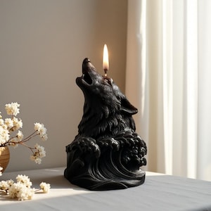May include: A black candle in the shape of a howling wolf, with a lit wick. The candle features detailed fur and sits on a wave design. The candle is on a table with white flowers in the background.