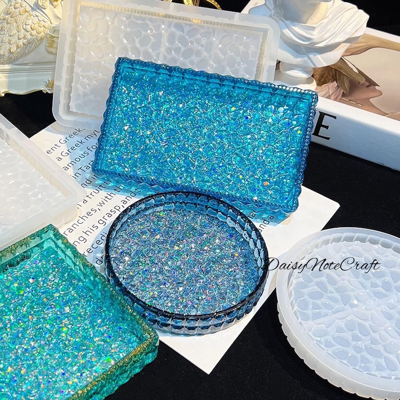May include: A collection of resin molds in various shapes, including rectangular trays and round coasters. The molds are clear and white, with some filled with sparkling blue glitter. The finished products have a textured, glittery appearance.