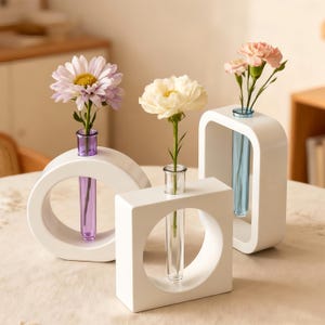 May include: Three white ceramic bud vases in different geometric shapes, each holding a single flower. The flowers are a light purple daisy, a white peony, and pink carnations. The vases have glass tubes.