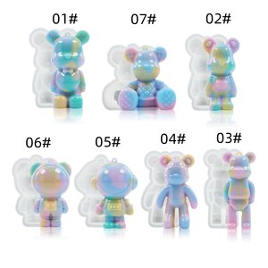 K&ouml;nnte beinhalten: Eine Sammlung farbenfroher B&auml;renfiguren aus Kunstharz in verschiedenen Posen. Die B&auml;ren sind haupts&auml;chlich in Pastellt&ouml;nen von Blau, Rosa und Gelb gehalten und haben ein gl&auml;nzendes Finish. Einige haben eine Schlaufe zum Aufh&auml;ngen. Das Bild zeigt einen Astronauten-B&auml;ren und einen sitzenden B&auml;ren.