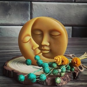 Puede incluir: Un jabón en forma de luna de color amarillo dorado con dos caras besándose, apoyado sobre una rodaja de madera. El jabón está decorado con detalles florales turquesa y naranja. El jabón tiene un diámetro de unos 10 cm.