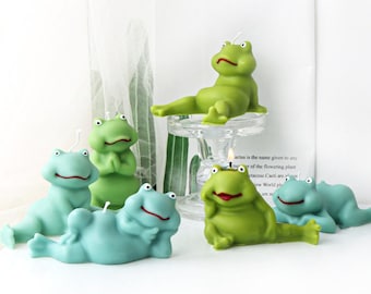 May include: A selection of frog-shaped candles in shades of green and blue. The candles are in various poses, including reclining and sitting. One candle is on a glass stand. The word "Cactus" is visible in the background.