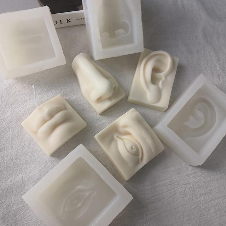 May include: A collection of off-white, sculpted soap bars and molds, each depicting a different facial feature: a nose, an ear, lips, and an eye. The soap bars are arranged on a light-colored surface, showcasing the detailed artistry of the human face.