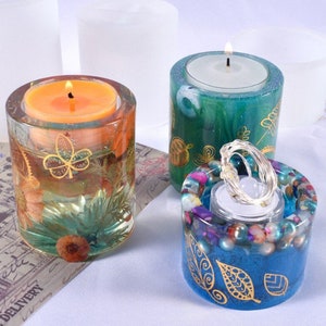 K&ouml;nnte beinhalten: Drei dekorative Kerzenhalter. Einer ist transparent mit eingebetteten Blumen und einem goldenen Blattdesign, das eine orangefarbene Kerze h&auml;lt. Ein weiterer ist gr&uuml;n mit goldenen Bl&auml;ttern. Der dritte ist blau mit goldenen Blattmustern und bunten Perlen.