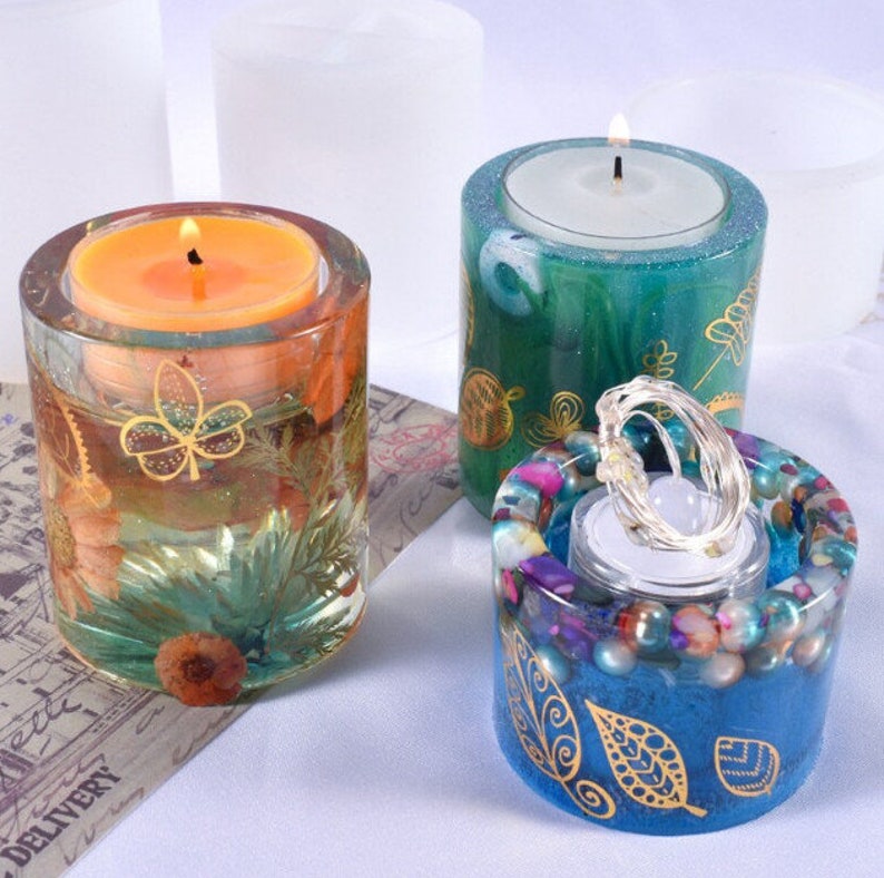 K&ouml;nnte beinhalten: Drei dekorative Kerzenhalter. Einer ist transparent mit eingebetteten Blumen und einem goldenen Blattdesign, das eine orangefarbene Kerze h&auml;lt. Ein weiterer ist gr&uuml;n mit goldenen Bl&auml;ttern. Der dritte ist blau mit goldenen Blattmustern und bunten Perlen.