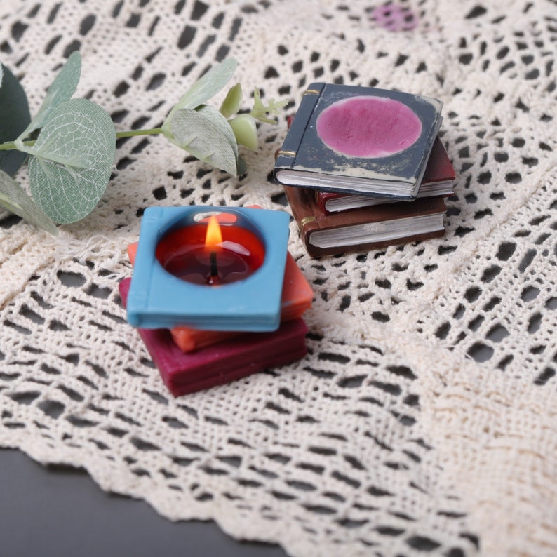 May include: A stack of miniature, colorful candles shaped like books and a candle with a burning flame. The candles are in shades of blue, red, and orange. The miniature books are brown and blue with gold accents. The items are arranged on a cream-colored, lace-like surface.