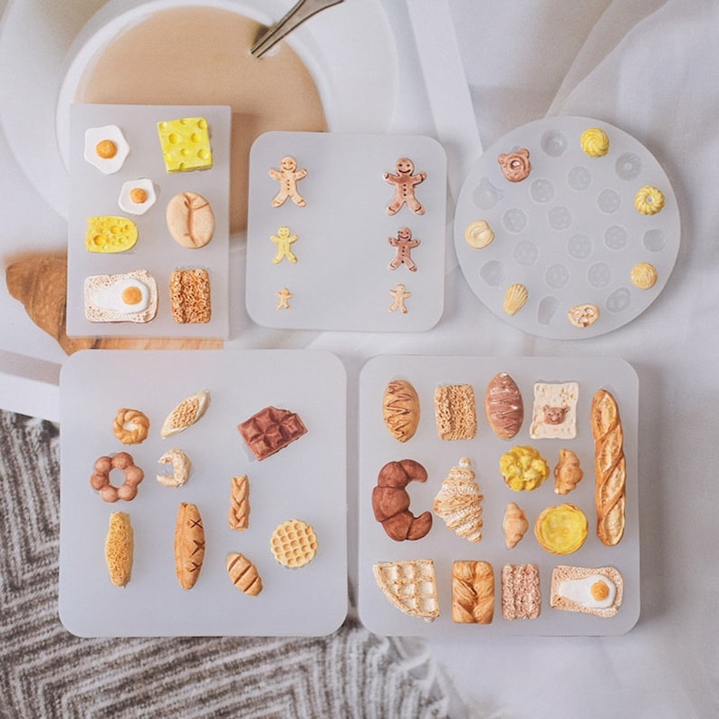 May include: A collection of silicone molds featuring miniature food designs. The molds include various baked goods like bread, croissants, and waffles, along with eggs, cheese, and gingerbread men. The molds are arranged on a light gray surface.