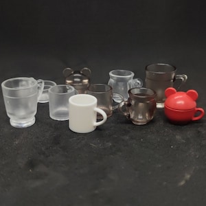 May include: A collection of miniature mugs in various styles and colors. The mugs are made of clear, frosted, and opaque materials, including a white mug and a red mug with a bear-shaped lid. The mugs are arranged on a dark surface.