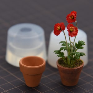 May include: A miniature terracotta flower pot and a matching silicone mold, alongside a second terracotta pot containing a small artificial plant with three vibrant red flowers, green leaves, and soil, all set against a dark grid background.
