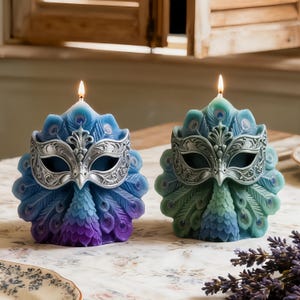 May include: Two peacock-shaped candles with lit wicks. One is blue and purple, the other is green and blue. Each candle features a silver-colored masquerade mask. The candles are on a floral patterned surface.