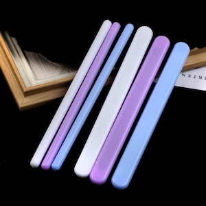 May include: An assortment of pastel-colored craft tools, including white, lavender, and blue sticks and flat tools. The tools are arranged diagonally against a dark background, suggesting a crafting or DIY theme.