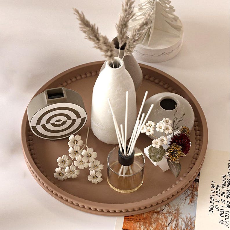 May include: A decorative tray with various home decor items. Includes a white vase with dried grass, a gray and white geometric object, a clear glass diffuser, and small floral arrangements. The tray is a light brown color.
