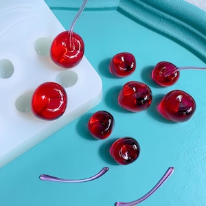 Può includere: Una collezione di ciliegie rosse traslucide in resina con gambi, alcune ancora attaccate a uno stampo bianco. Le ciliegie sono sparse su una superficie turchese, suggerendo un progetto fai-da-te. La resina ha una finitura lucida.