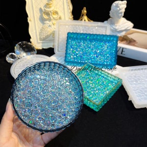 May include: A collection of resin art pieces in various shapes, including a round coaster and rectangular trays, all featuring a sparkling blue glitter design. The pieces are displayed on a dark surface, with a frame and decorative objects in the background.