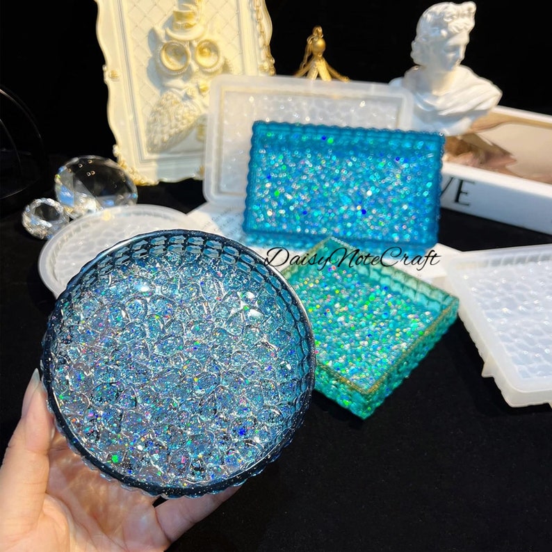 May include: A collection of resin art pieces in various shapes, including a round coaster and rectangular trays, all featuring a sparkling blue glitter design. The pieces are displayed on a dark surface, with a frame and decorative objects in the background.