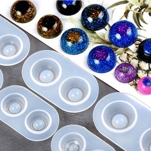 May include: A collection of resin art supplies, including clear silicone molds with spherical cavities and various colorful, glitter-filled resin spheres in shades of blue, purple, and gold. The molds are light blue.