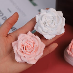 Pode incluir: Duas rosas de papel, uma branca e outra rosa claro, são seguradas em uma mão. As rosas exibem arranjos intrincados de pétalas, dando uma aparência delicada e artesanal. O fundo está desfocado, atraindo a atenção para as flores.