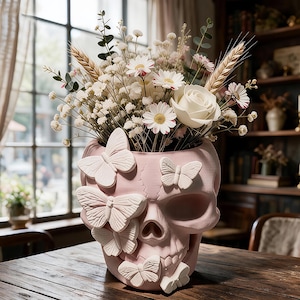 May include: A light pink skull-shaped vase adorned with several raised white butterfly figures. The vase is filled with a bouquet of white daisies, a single white rose, dried wheat stalks, and small sprigs of green eucalyptus foliage.