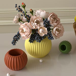 Può includere: Una composizione floreale in un vaso giallo scanalato con rose rosa chiaro e bacche blu scuro. Sono presenti anche una ciotola scanalata color terracotta più piccola e un oggetto verde scanalato. Perle sparse aggiungono un tocco decorativo.