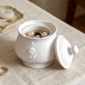 Peut inclure: Un pot en céramique blanche avec un bord festonné et un couvercle assorti, présentant un relief de style camée sur le devant. Le pot contient diverses bagues en perles et dorées, tandis qu'une bague dorée avec des pierres repose à côté.