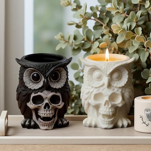 May include: Two owl-shaped candle holders, one black and one white, each featuring a skull integrated into the chest area. The white owl holder contains a lit candle with a warm yellow flame. Both figures have detailed feathers and wide eyes.