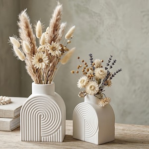May include: Two white ceramic vases featuring a textured spiral arch pattern on the front. The taller vase contains a bouquet of beige pampas grass and white dried flowers. The shorter vase holds a mix of dried white flowers and purple lavender stems.