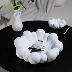 Puede incluir: Una bandeja decorativa blanca en forma de nube contiene un frasco de perfume, llaves del coche y joyas. Una bandeja más pequeña a juego y un jarrón blanco con vegetación también están sobre la superficie negra. Los objetos están colocados sobre una mesa negra.