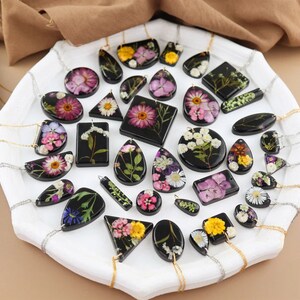 May include: A selection of resin pendants with dried flowers in diverse shapes, including circles, squares, and teardrops. The pendants are on silver chains and presented on a white tray. The flowers display shades of pink, yellow, and white.