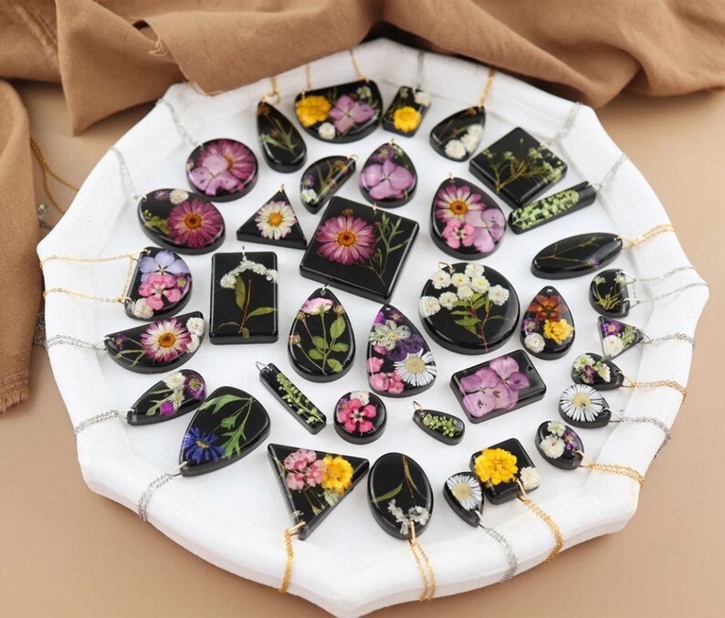 May include: A selection of resin pendants with dried flowers in diverse shapes, including circles, squares, and teardrops. The pendants are on silver chains and presented on a white tray. The flowers display shades of pink, yellow, and white.