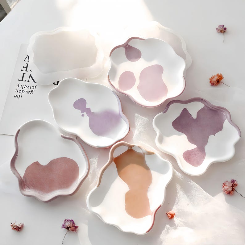 May include: A collection of white ceramic plates with irregular, wavy edges. Each plate features a unique abstract design in shades of pink, purple, and brown. The plates are arranged on a white surface, with a few dried flowers scattered around.
