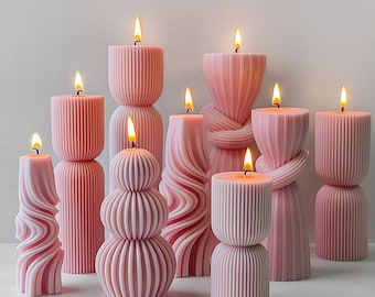 May include: A collection of pink pillar candles with various designs, including ribbed, twisted, and stacked shapes. Each candle is lit, with a bright yellow flame. The candles are arranged on a white surface, creating a soft, warm ambiance.