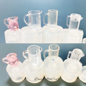May include: A selection of miniature clear and pink glass jugs and mugs. The jugs have handles and spouts, while the mugs are cylindrical. Some have lids. The pink jug features a floral design. The items are displayed on white cylindrical bases.