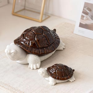 May include: Two ceramic turtle figurines. The larger turtle has a dark brown shell and a white body. The smaller turtle has a similar design. The figurines are decorative and likely for home decor.