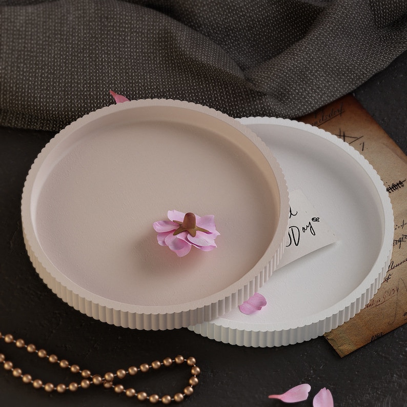 Pu&ograve; includere: Due vassoi rotondi con bordi scanalati, uno rosa chiaro e l'altro bianco. Un fiore rosa &egrave; appoggiato sul vassoio rosa. Sono visibili anche una collana di perline dorate e un biglietto con "Good Day". I vassoi sono probabilmente decorativi.