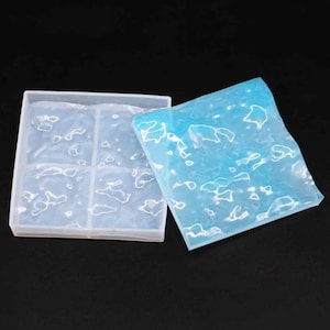 May include: A clear, square silicone mold with six compartments, next to a blue, textured silicone sheet. The mold and sheet have a wavy, organic pattern. The items are on a black background.