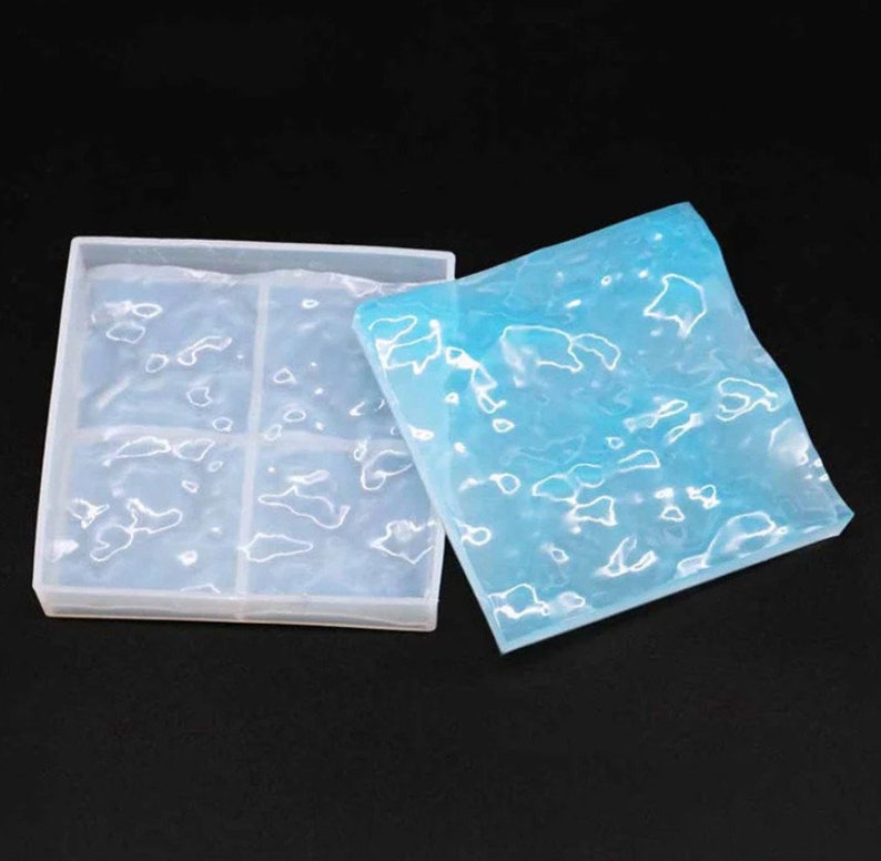 May include: A clear, square silicone mold with six compartments, next to a blue, textured silicone sheet. The mold and sheet have a wavy, organic pattern. The items are on a black background.