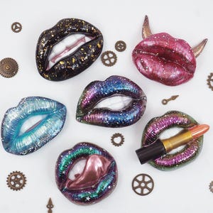 Puede incluir: Una variedad de objetos decorativos con forma de labios en varios colores, incluyendo negro con motas doradas, azul y morado, con un labial y pequeños engranajes dispersos. Los labios tienen un acabado brillante y están sobre un fondo blanco.