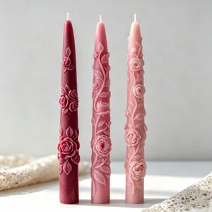 Peut inclure: Trois bougies effilées dans des tons bordeaux profond, vieux rose et rose clair. Chaque bougie présente des motifs floraux en relief complexes de roses et de vignes s'enroulant autour de l'extérieur, avec une mèche au sommet de chacune.