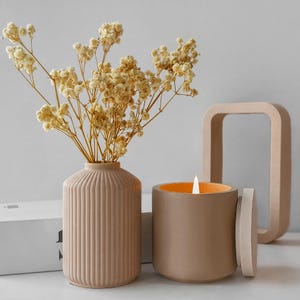 Pode incluir: Uma vela de cerâmica bege com um pavio aceso e uma tampa a condizer está ao lado de um vaso canelado com flores secas. Um objeto decorativo quadrado castanho claro está no fundo. A vela tem um interior laranja.