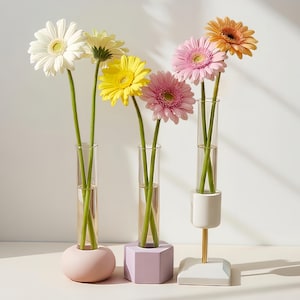 May include: Gerbera daisies in clear glass vases with pastel-coloured bases. The flowers are white, yellow, pink, and orange. The vases are arranged on a white surface, with soft light and shadow.
