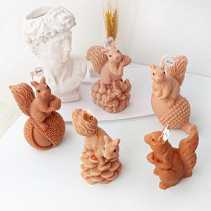 May include: A collection of brown squirrel-shaped candles, some perched on pinecones and nuts. A white bust of a classical figure and a sprig of wheat are in the background. Each candle has a small tag that says "Smile".