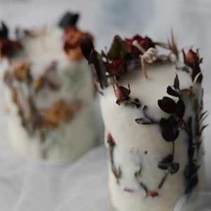 May include: Two white pillar candles are adorned with dried flowers and foliage. The candles have a rustic, natural aesthetic, with the dried elements adding texture and color. The candles are a soft, off-white color.