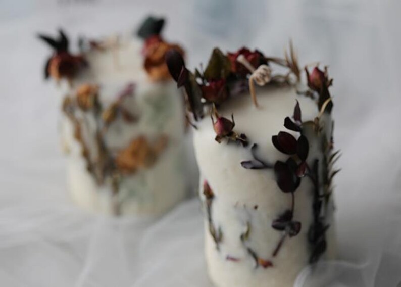 May include: Two white pillar candles are adorned with dried flowers and foliage. The candles have a rustic, natural aesthetic, with the dried elements adding texture and color. The candles are a soft, off-white color.