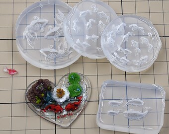 May include: A collection of clear resin molds featuring aquatic themes. The molds include circular and heart-shaped designs with goldfish and aquatic plants. A small, red and white fish figurine is also present.