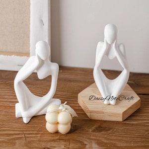 Puede incluir: Dos esculturas blancas abstractas de figuras humanas, una sentada y otra de pie sobre una base de madera, junto con una pequeña vela redonda de color crema. Esculturas minimalistas y modernas.