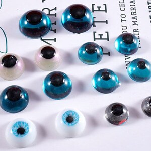May include: A collection of glass eye cabochons in various sizes and colors. The eyes feature a dark pupil and come in shades of blue, white, and iridescent hues. Some eyes have a clear, domed shape, while others are more opaque.