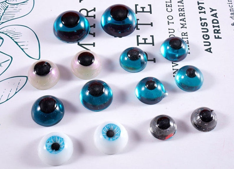 May include: A collection of glass eye cabochons in various sizes and colors. The eyes feature a dark pupil and come in shades of blue, white, and iridescent hues. Some eyes have a clear, domed shape, while others are more opaque.